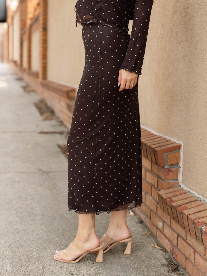A woman wearing a black midi skirt with white polka dots and a matching long-sleeve top, paired with clear strap heeled sandals, standing on a sidewalk next to a brick wall.