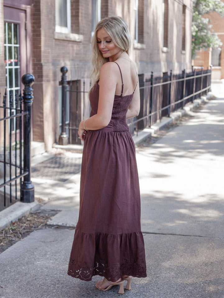 A woman wearing a sleeveless, tiered maxi dress in a dusty plum-brown color, standing on a city sidewalk next to a brick building.