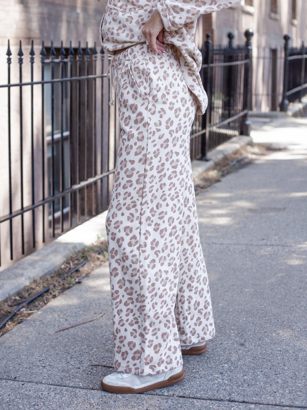 A person standing on a city sidewalk wearing loose, wide-leg pants in a beige and brown leopard print, paired with a matching top and white slip-on shoes.