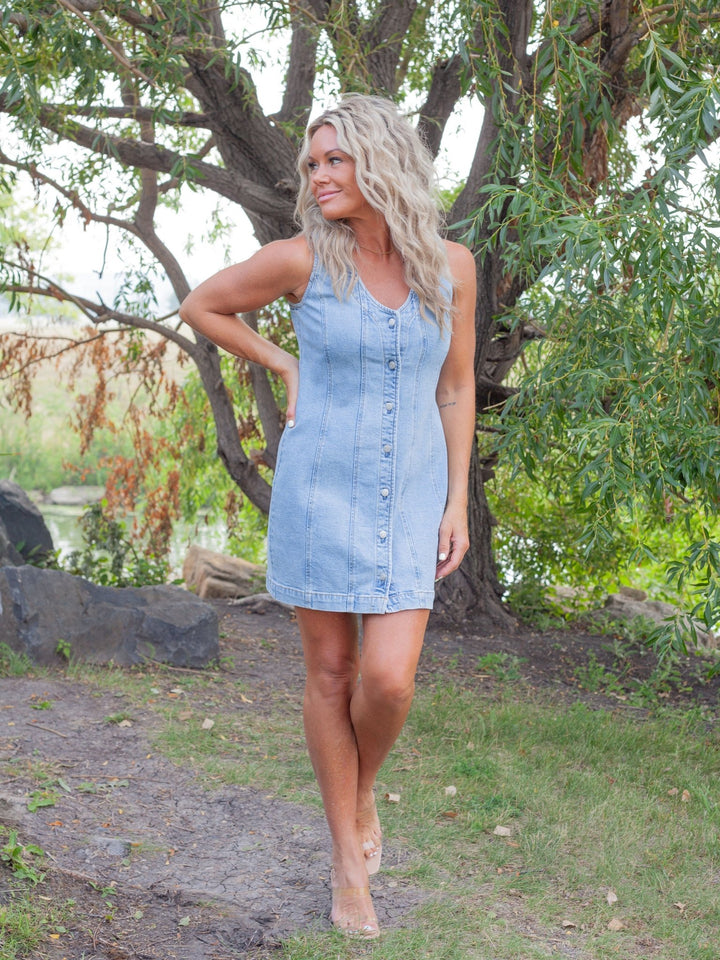 A woman modeling a light blue denim mini dress with a button-down front, photographed outdoors in a park setting with trees and grass.