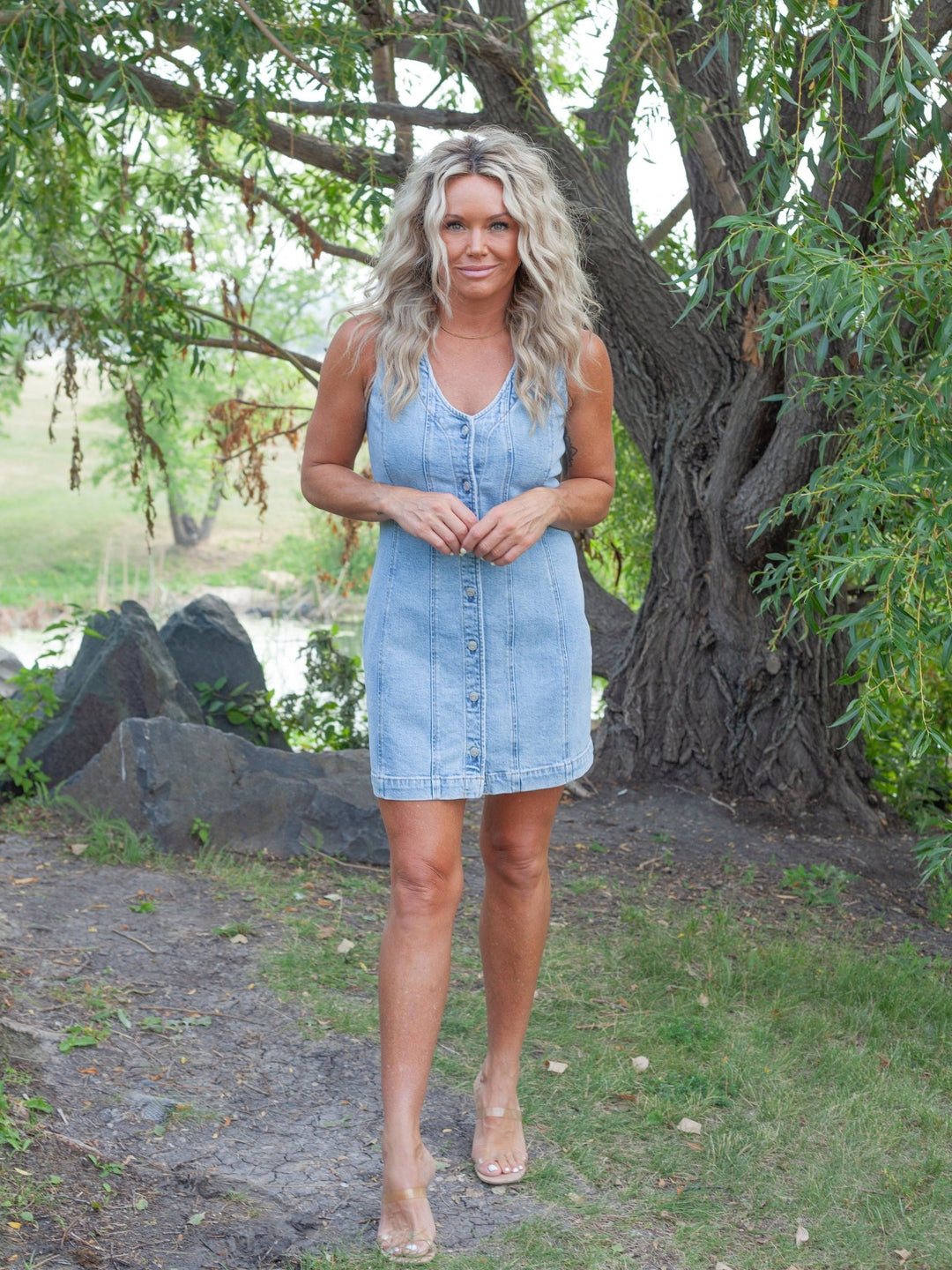 A woman wearing a light blue denim mini dress with a button-down front, standing outdoors in a park-like setting with trees and rocks in the background.