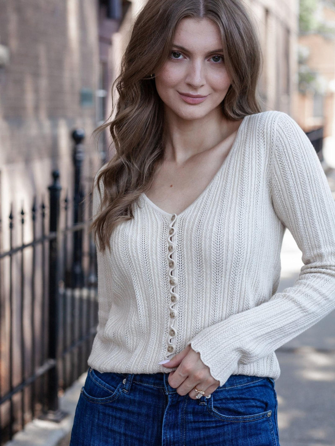 A woman wearing a cream-colored ribbed knit cardigan with a button-down front and blue jeans, standing outdoors on a city street.
