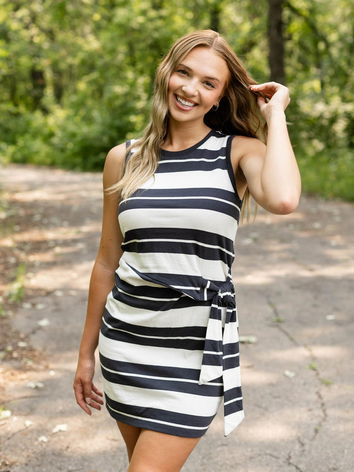 A woman wearing a sleeveless black and white horizontally striped mini dress with a tie waist, standing outdoors on a paved path with a blurred green forest background.