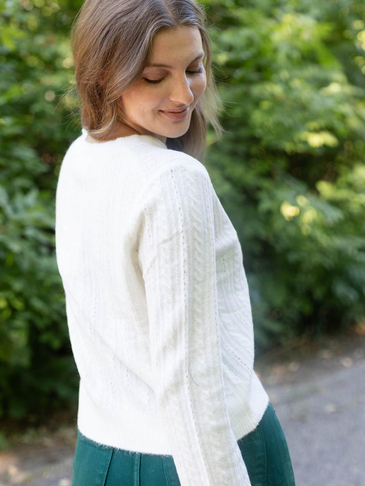 A woman wearing a white cable-knit cropped sweater and a dark green skirt, photographed outdoors against a blurred background of green foliage.