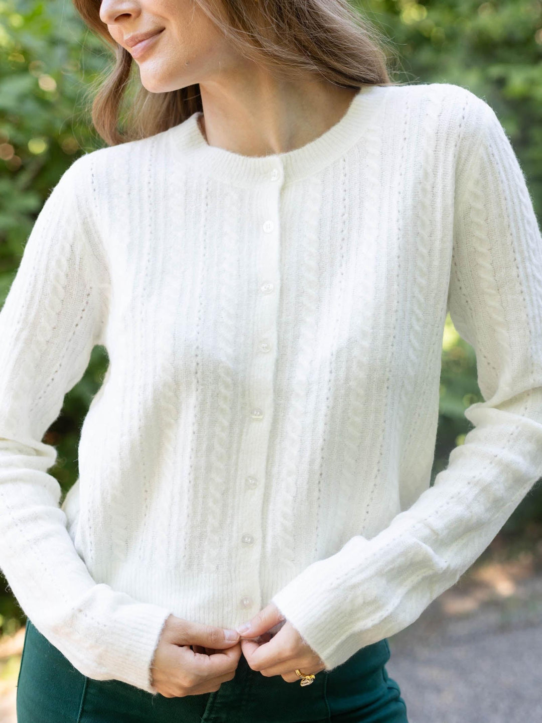 A woman wearing a cream cable-knit button-front cardigan sweater, shown from the chest down in an outdoor setting with blurred green foliage in the background.