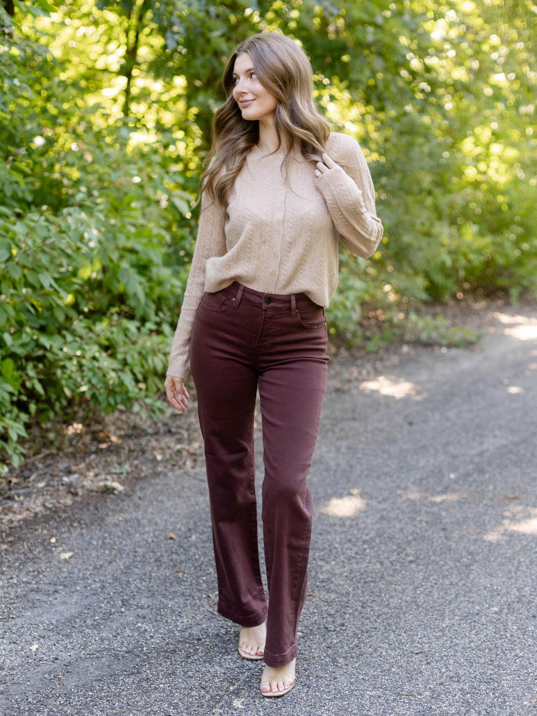 A woman walking on a gravel path surrounded by green trees, wearing a beige knit sweater and dark burgundy flare jeans.