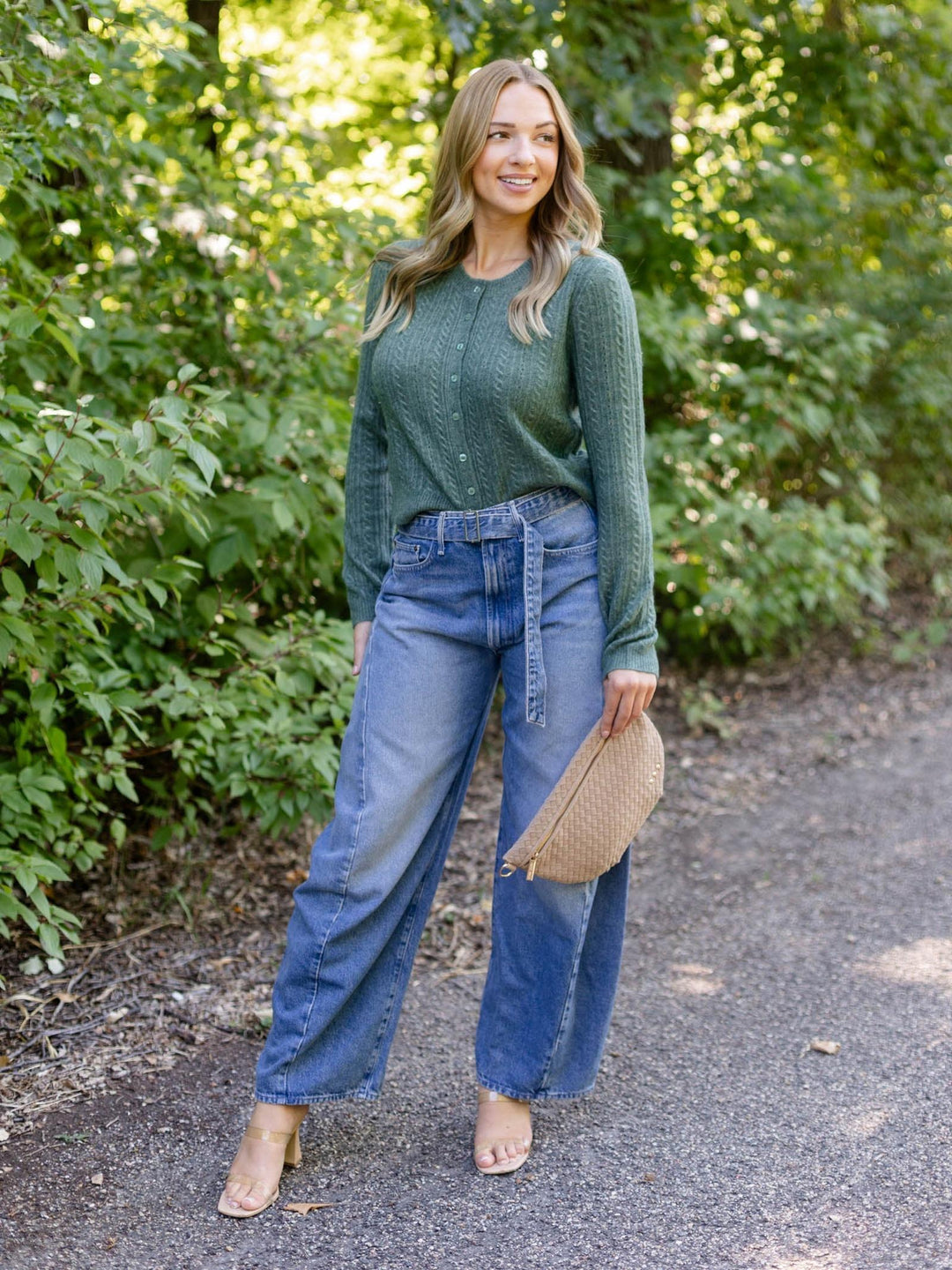 A woman stands on a gravel path surrounded by green foliage, wearing a green knit long-sleeve top, wide-leg blue jeans, and holding a woven straw hat.