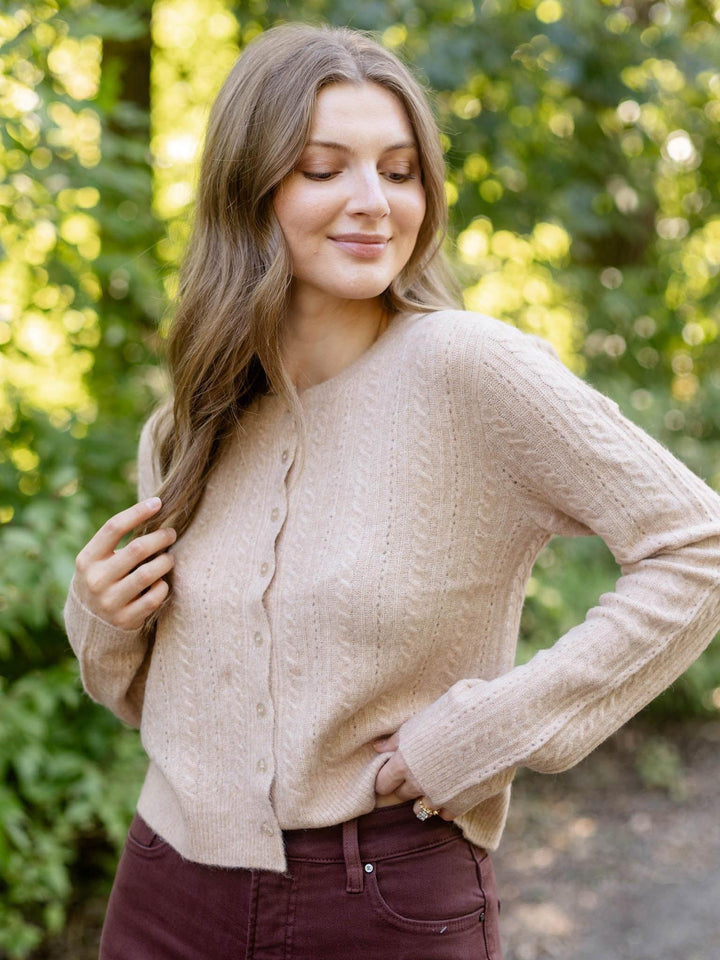 A woman wearing a beige cable-knit cardigan sweater and dark burgundy pants, standing outdoors in front of a blurred green forest background.