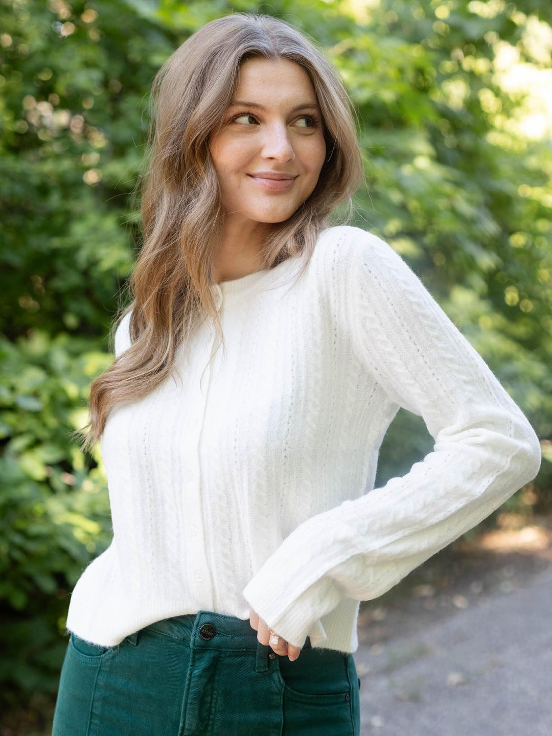 A woman wearing a white cable-knit long-sleeve sweater and dark green pants, standing outdoors in front of a blurred green foliage background.