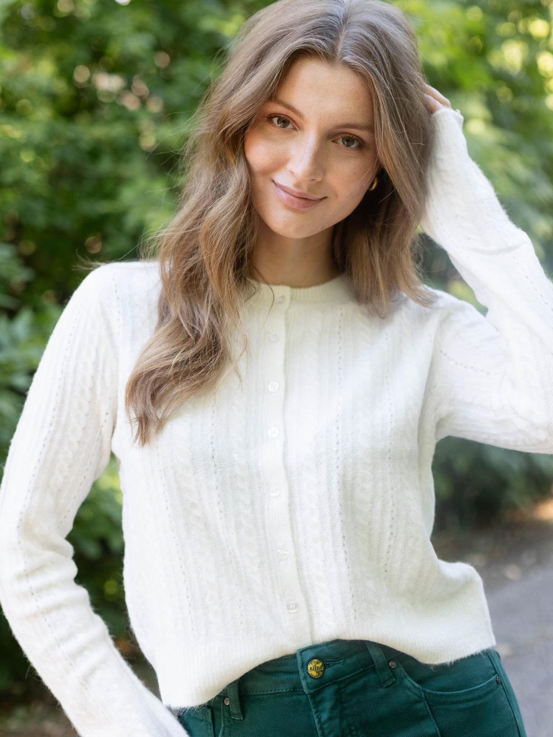 A woman with long wavy light brown hair wearing a white cable-knit button-up sweater and dark green jeans, posing outdoors with a blurred green foliage background.