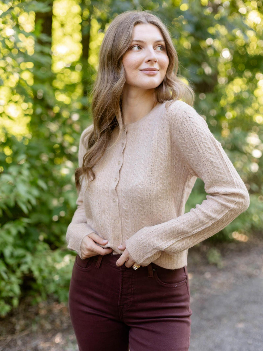 A woman wearing a beige cable-knit cardigan and burgundy pants, standing outdoors in a sunlit forest setting.