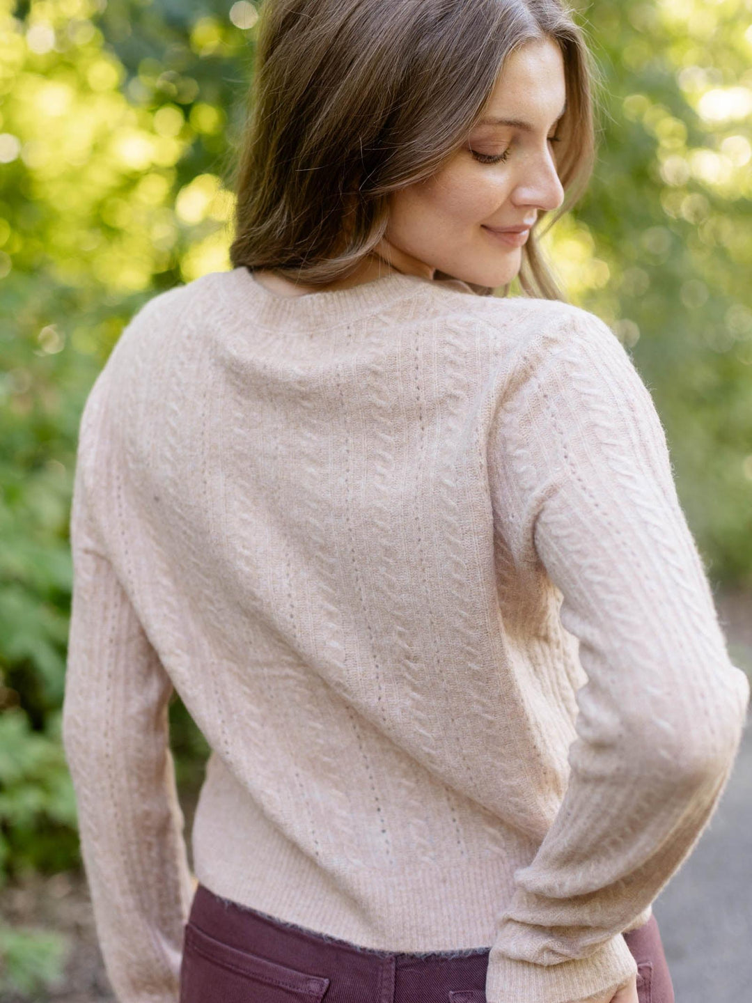 A woman wearing a light beige cable-knit sweater, photographed from behind in an outdoor setting with a blurred green background.