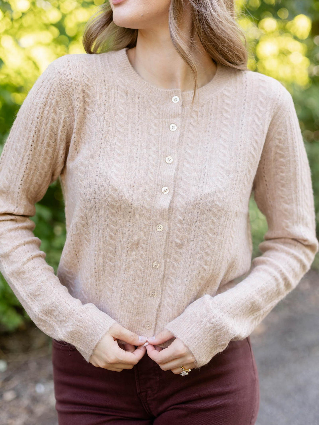A woman wearing a beige cable-knit button-front cardigan sweater, paired with burgundy pants, photographed outdoors with a blurred green background.