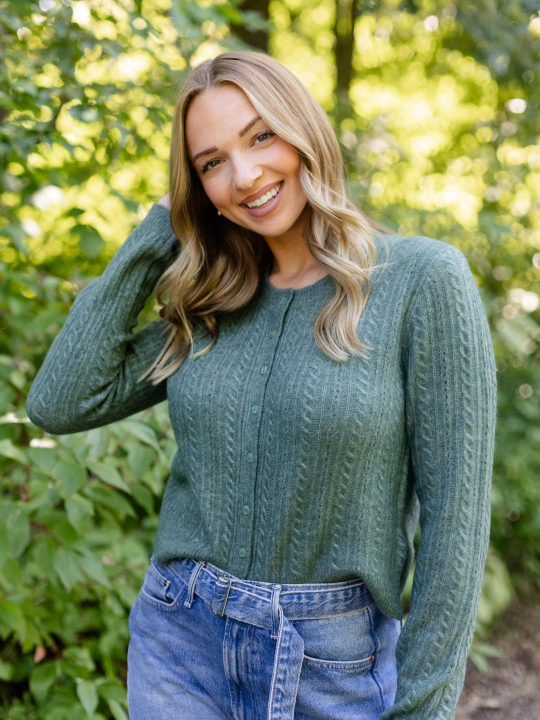 A smiling blonde woman wearing a sage green cable-knit sweater and blue jeans, posing outdoors against a soft-focus background of green foliage.