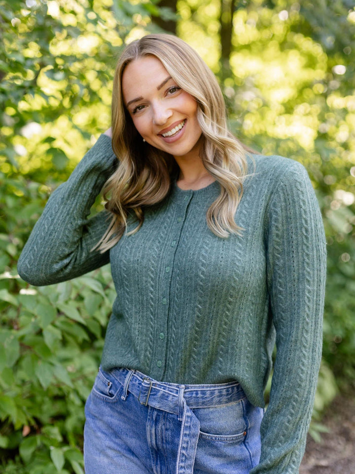 A smiling blonde woman wearing a sage green cable-knit sweater and blue jeans, posing outdoors against a soft-focus background of green foliage.