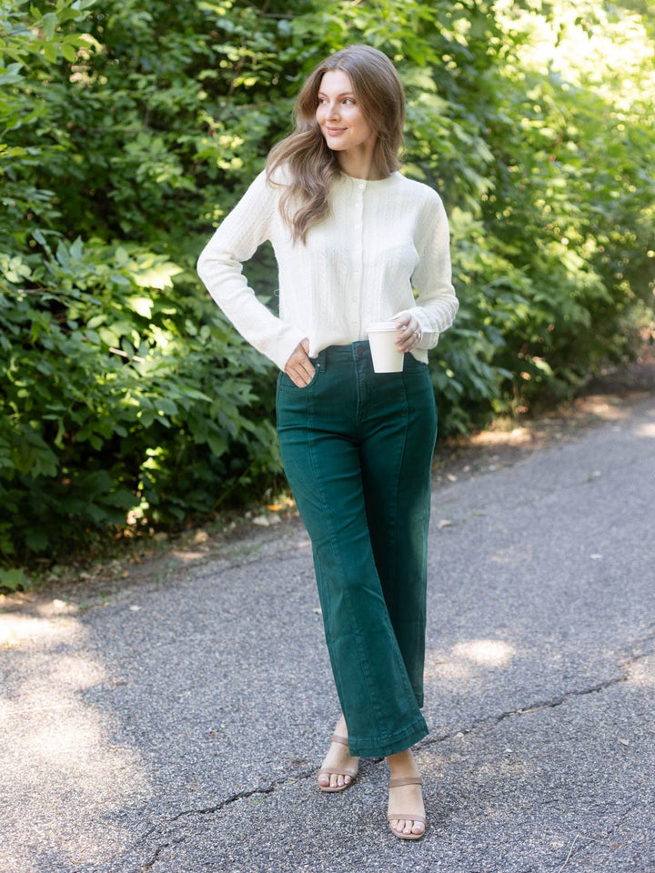 A woman stands on a paved path outdoors wearing a cream ribbed mock-neck long-sleeve top and dark green wide-leg pants, with beige heeled sandals.