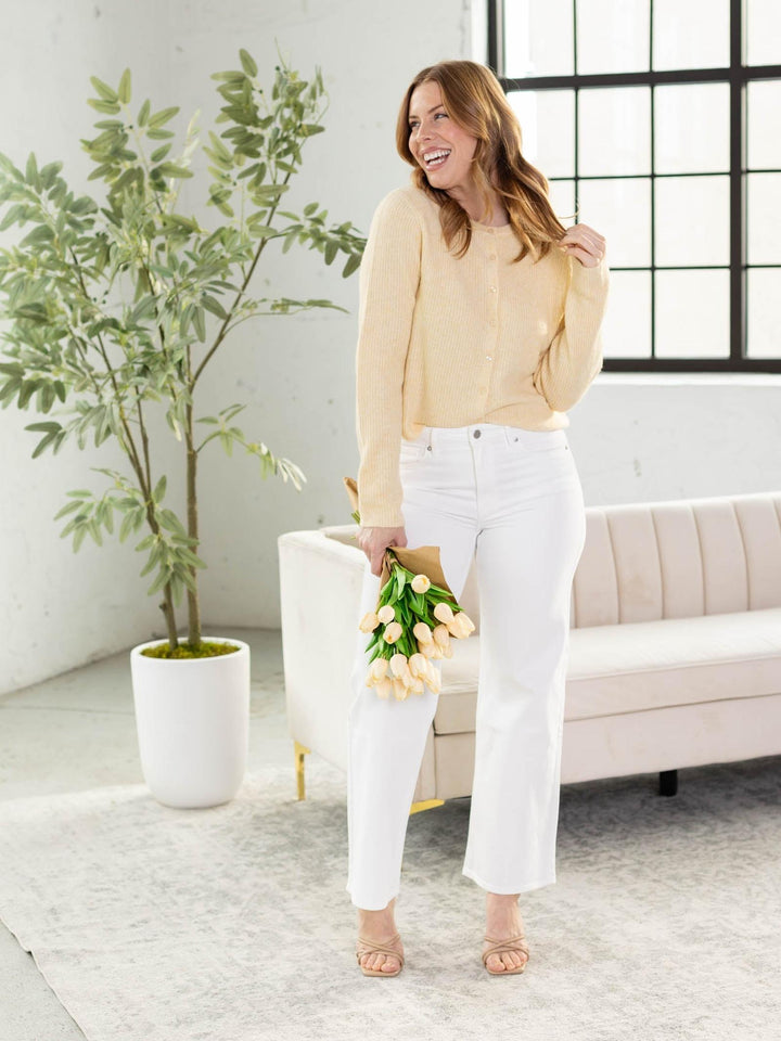 Woman in a yellow blouse and white pants standing in a bright room with a plant and sofa.