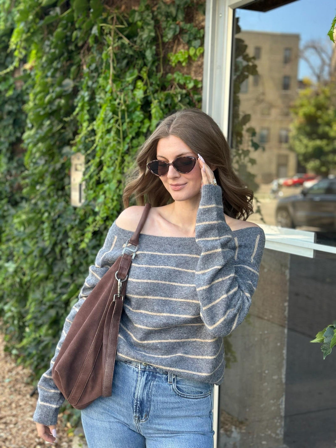 A woman wearing a gray and white striped off-the-shoulder sweater, blue jeans, and sunglasses, carrying a brown shoulder bag while standing outdoors next to a window with ivy-covered walls.