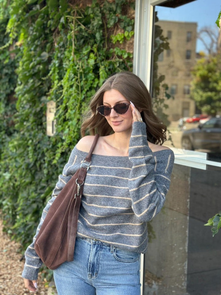 A woman wearing a gray and white striped off-the-shoulder sweater, blue jeans, and sunglasses, carrying a brown shoulder bag while standing outdoors next to a window with ivy-covered walls.