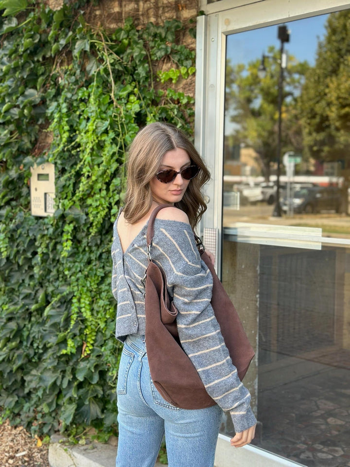 A woman wearing a gray and white striped off-the-shoulder sweater, blue jeans, and sunglasses, carrying a brown suede shoulder bag. She is standing outdoors next to a brick wall covered in green ivy and a glass door.
