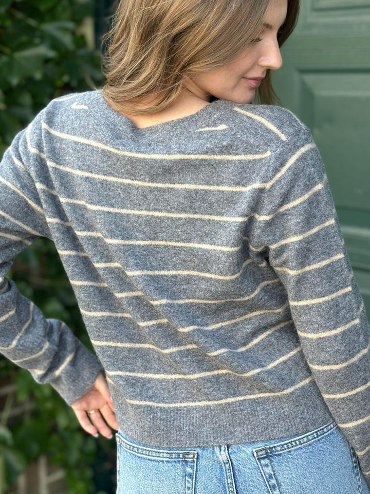 A woman wearing a gray knit sweater with thin cream horizontal stripes, paired with light blue jeans, photographed outdoors against a green door and foliage.