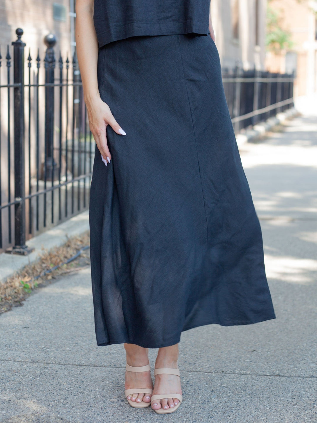 A person wearing a long, flowing black midi skirt with a side slit, paired with beige strappy heeled sandals, standing on a paved sidewalk next to a black metal fence.