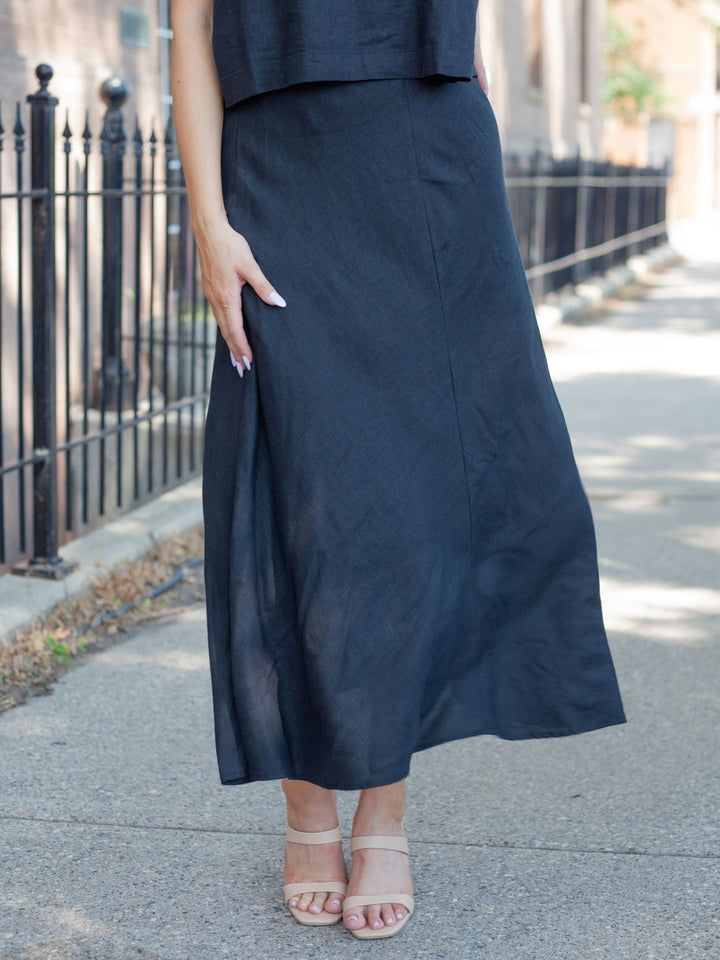 A person wearing a long, flowing black midi skirt with a side slit, paired with beige strappy heeled sandals, standing on a paved sidewalk next to a black metal fence.