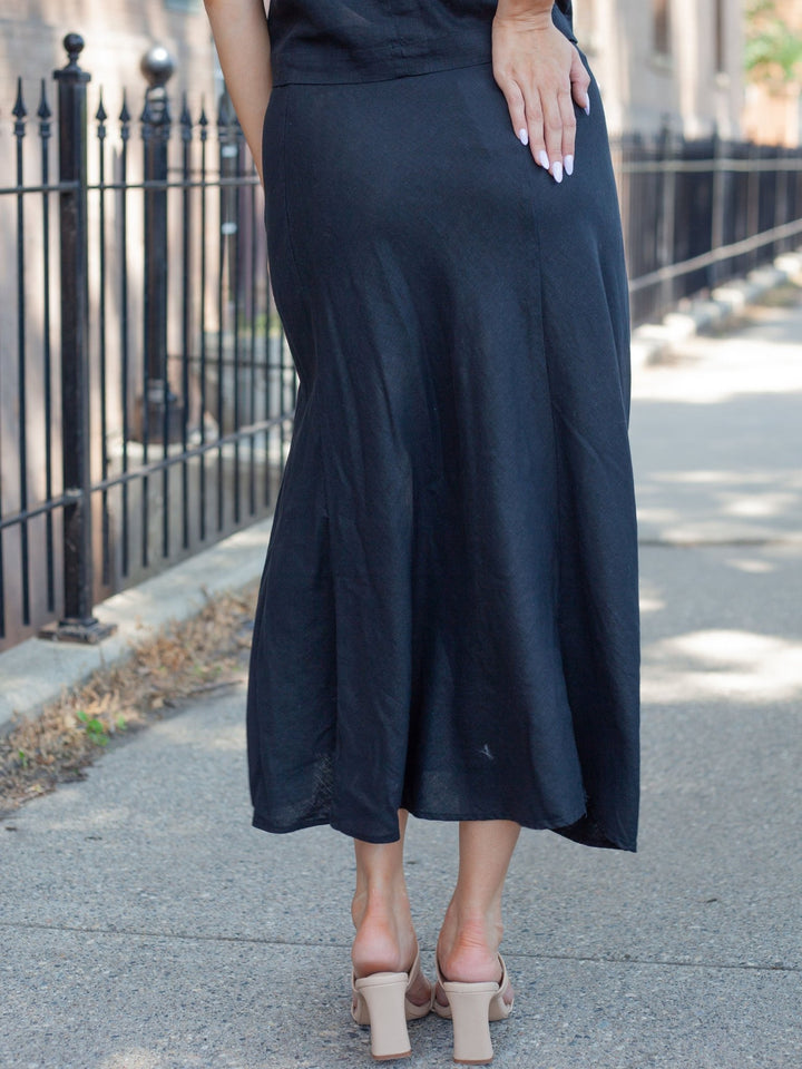 A woman wearing a flowing black midi skirt with a slight flare, paired with beige block-heel sandals, standing on a sidewalk next to a black metal fence.