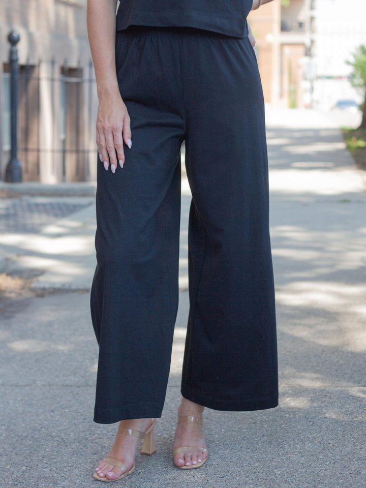 A person wearing black wide-leg cropped pants with a matching black top, standing outdoors on a paved street.