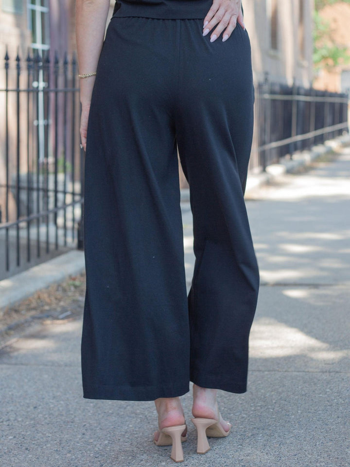 Black wide-leg cropped pants shown from the back on a model walking outdoors on a sidewalk.