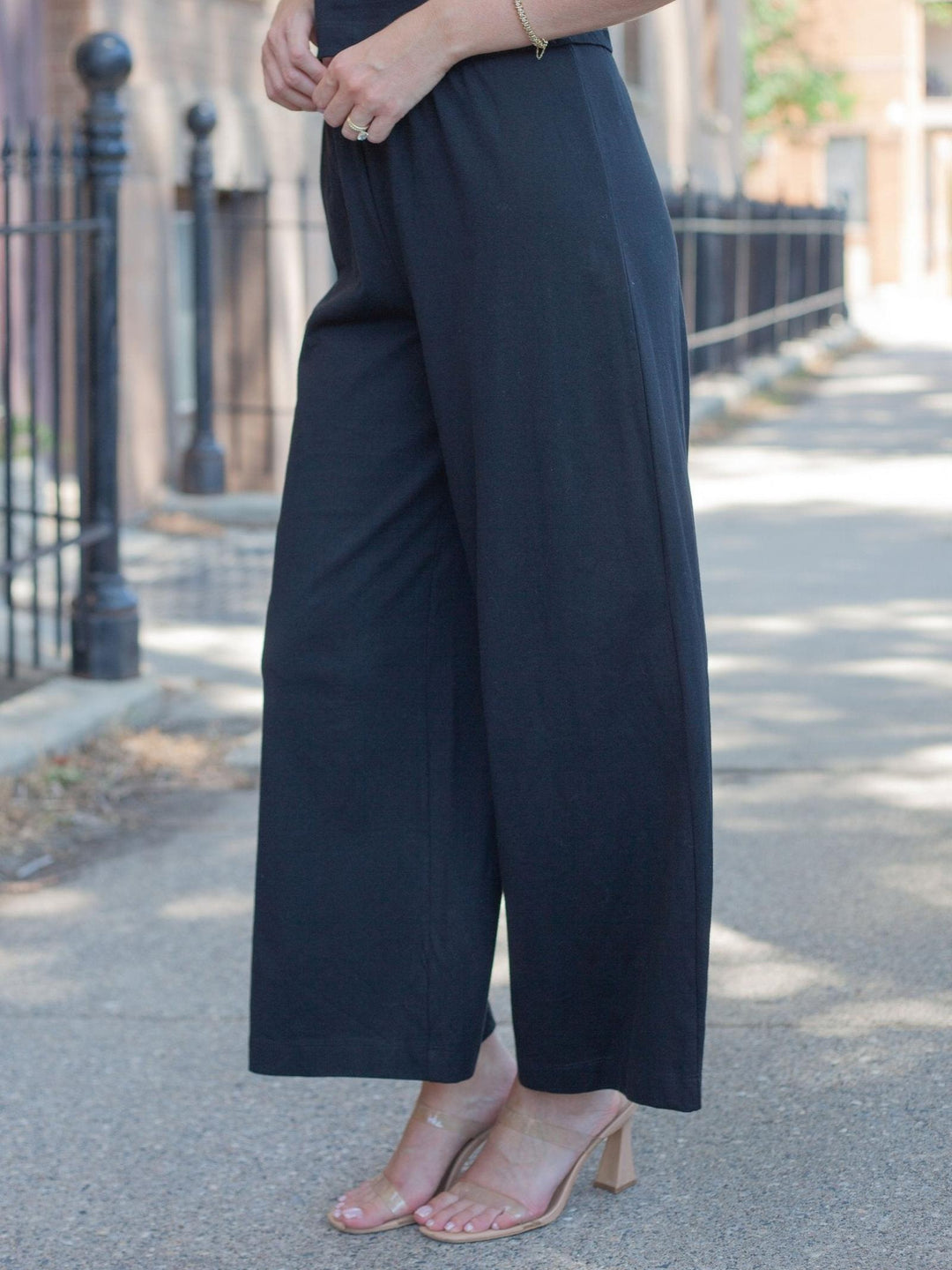 A person wearing loose, wide-leg black pants and clear strap heels standing on a sunlit sidewalk outdoors.