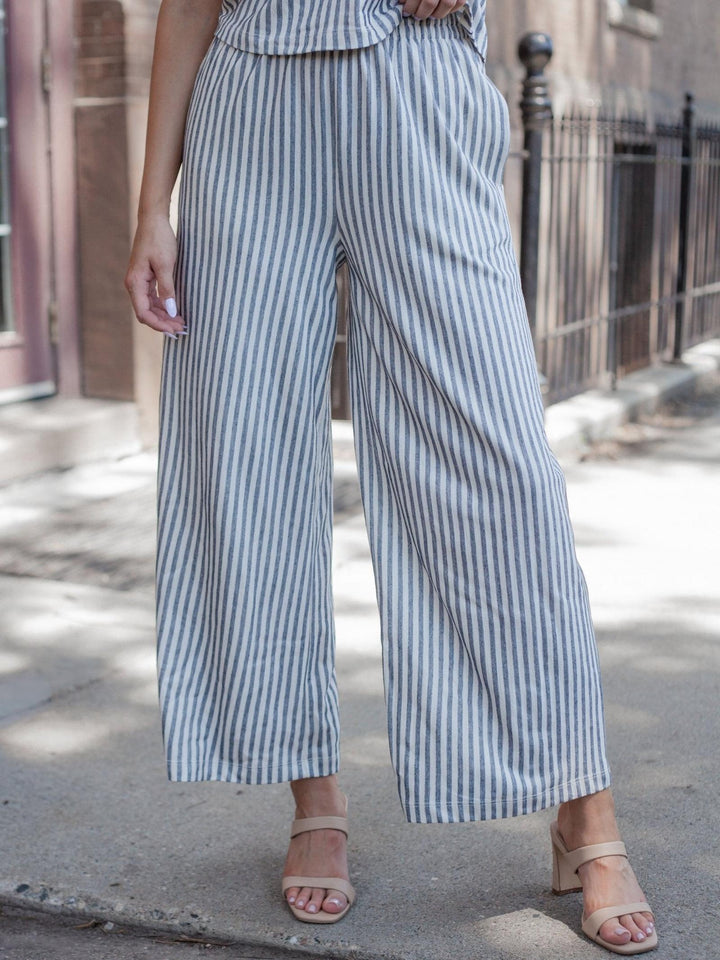 A person wearing wide-leg cropped pants with vertical blue and white stripes, paired with beige heeled sandals, standing outdoors on a sidewalk.
