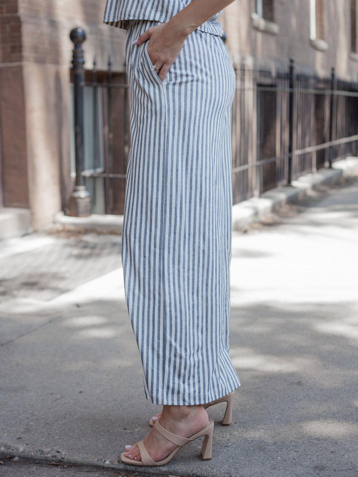 A person wearing a matching blue and white vertically striped two-piece set with wide-leg pants and a cropped top, paired with beige heeled sandals, standing on a city sidewalk.