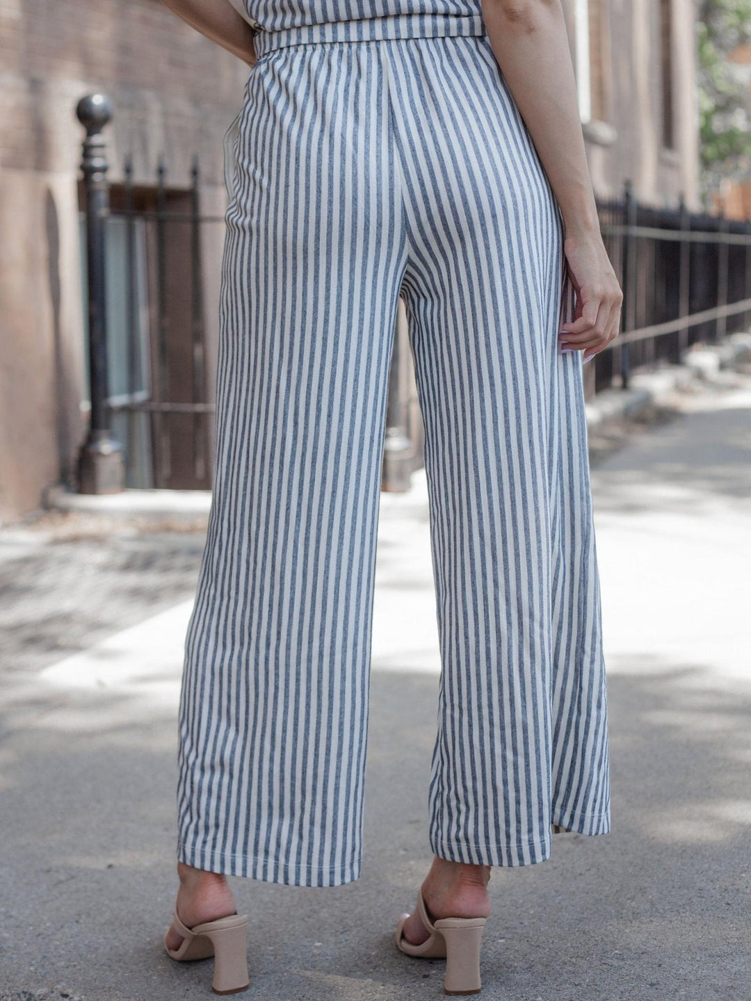 A person shown from the back wearing high-waisted, wide-leg striped pants in blue and white, paired with beige block-heel mule sandals on an outdoor city sidewalk.