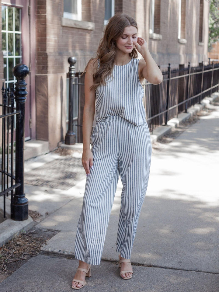 A woman stands on a city sidewalk wearing a sleeveless jumpsuit with vertical blue and white stripes, paired with nude heeled sandals.