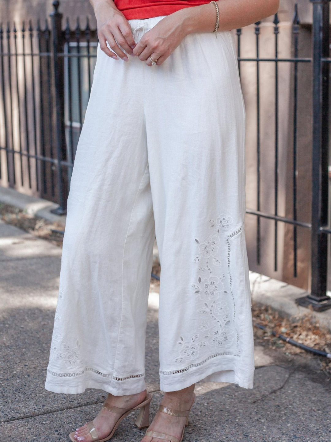 White wide-leg cropped pants with eyelet embroidery near the hem, shown on a model outdoors in front of a black metal fence.
