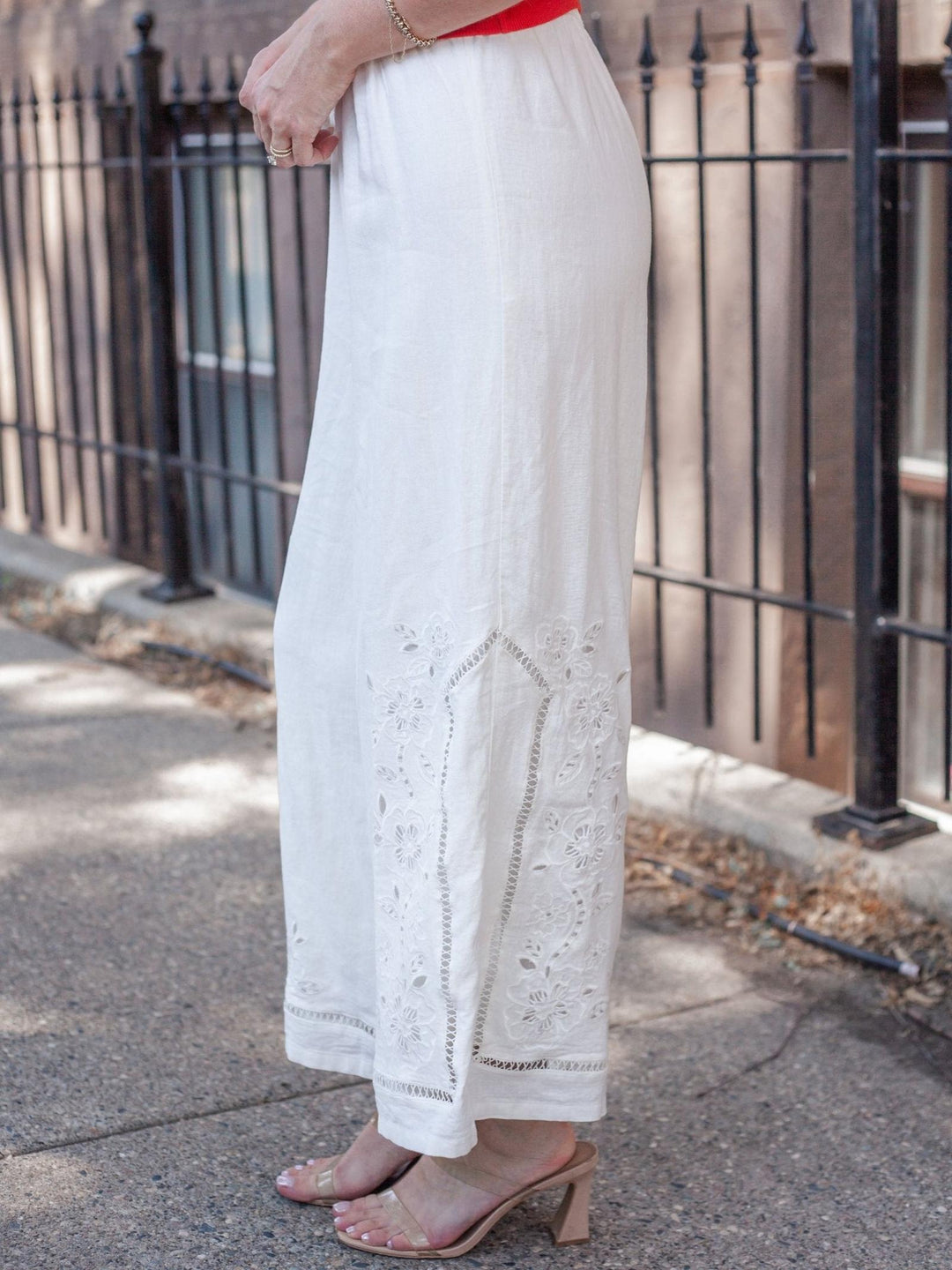 A person wearing a white wide-leg skirt with floral eyelet embroidery near the hem, paired with beige heeled sandals, standing outdoors on a sidewalk beside a black metal fence.