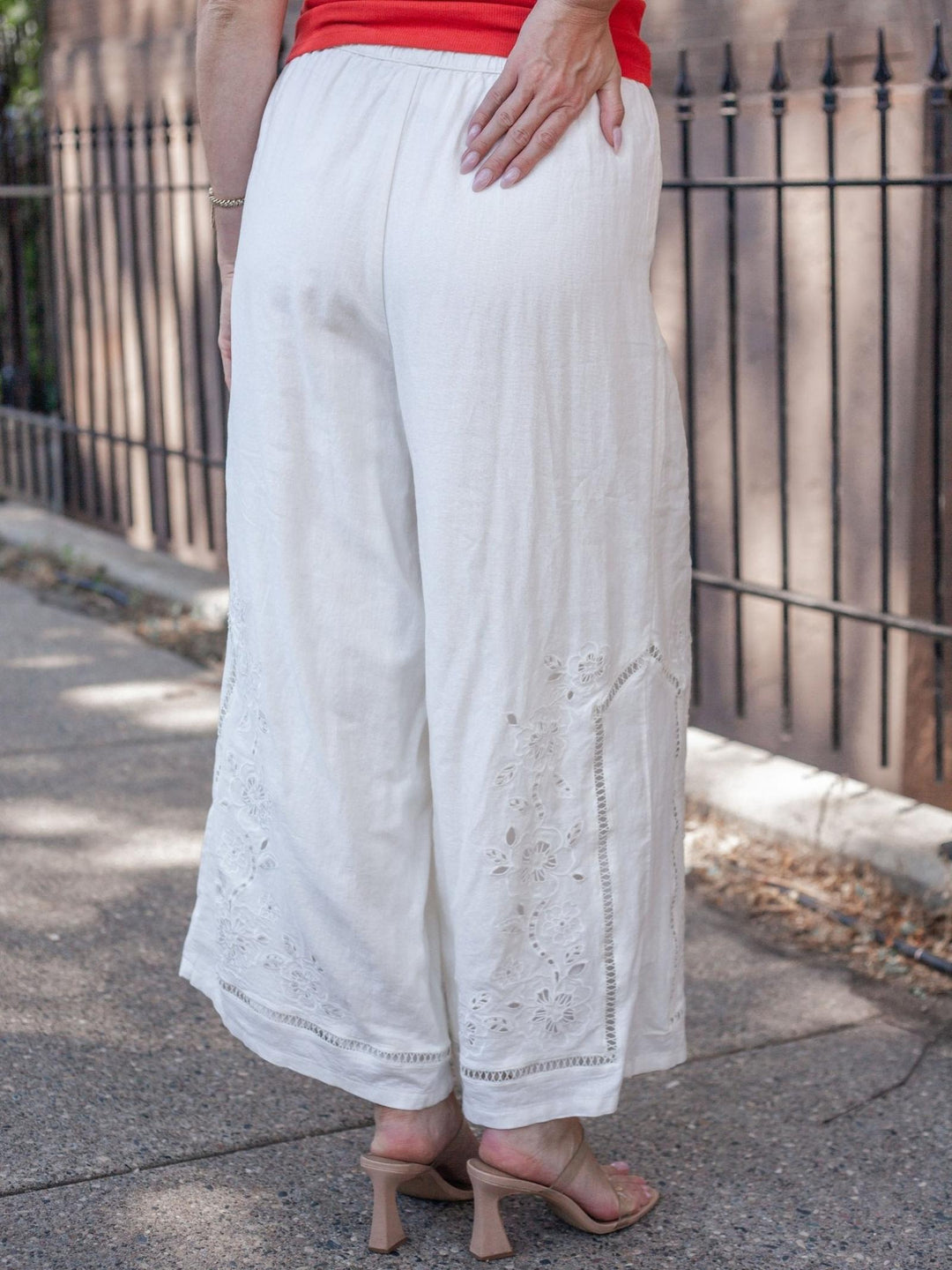 White wide-leg cropped pants with eyelet embroidery near the hems, shown on a model from the back wearing beige heeled sandals on a sidewalk.