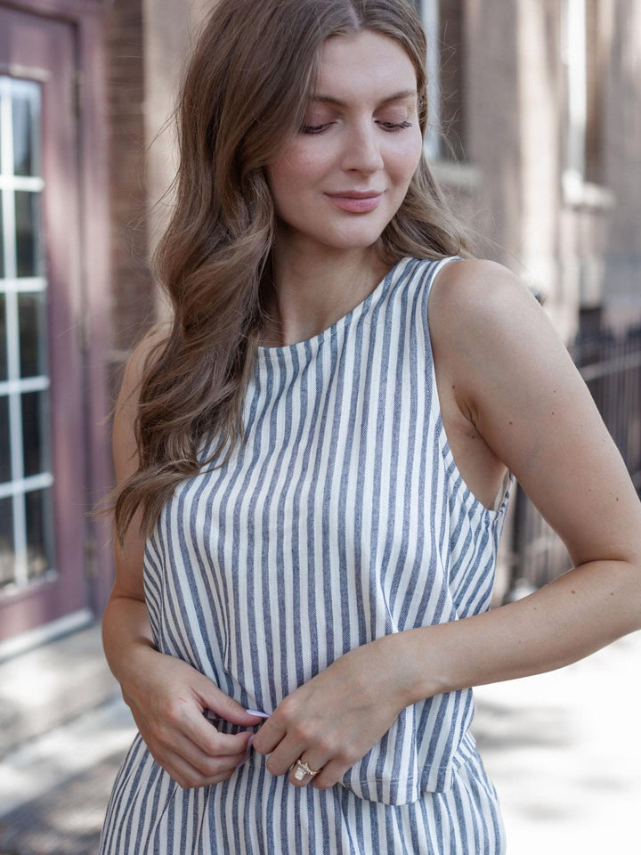 A woman wearing a sleeveless top with vertical navy and white stripes, standing outdoors in front of a blurred brick building.