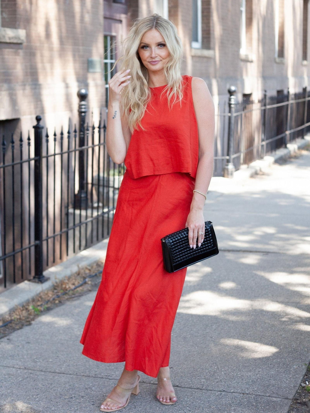 A woman stands on a city sidewalk wearing a sleeveless red two-piece outfit consisting of a cropped top and a long skirt, holding a black clutch bag.