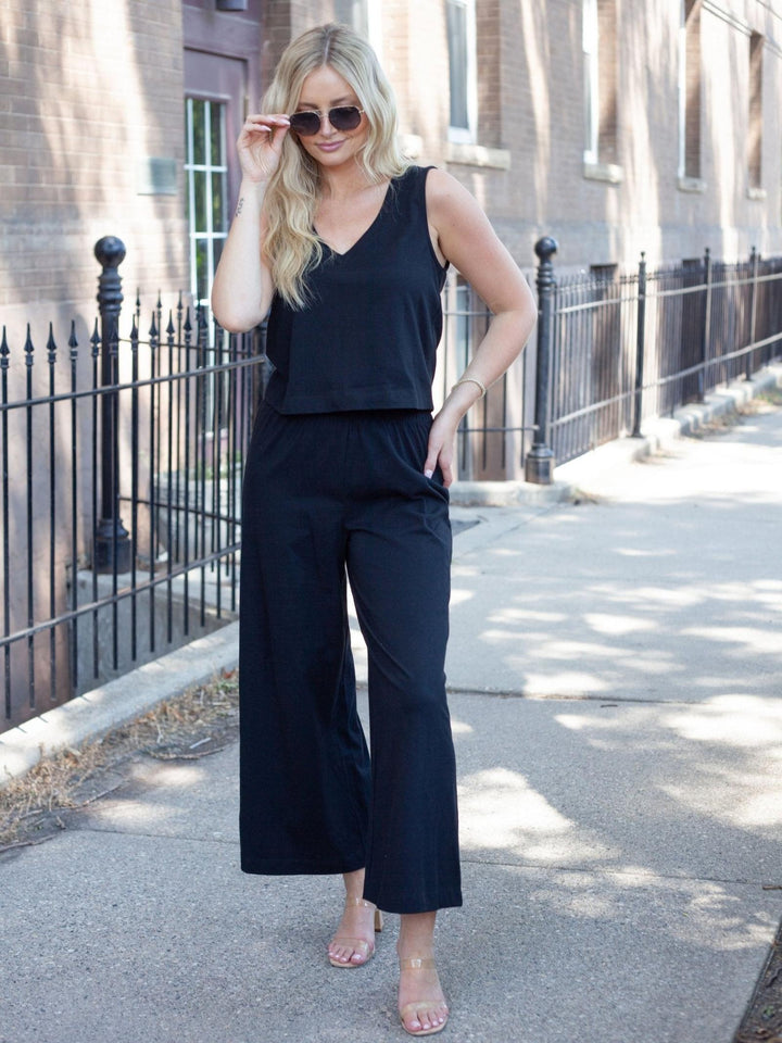 A woman stands on a city sidewalk wearing a black sleeveless V-neck top and matching black wide-leg cropped pants, with nude high-heel sandals and dark sunglasses.