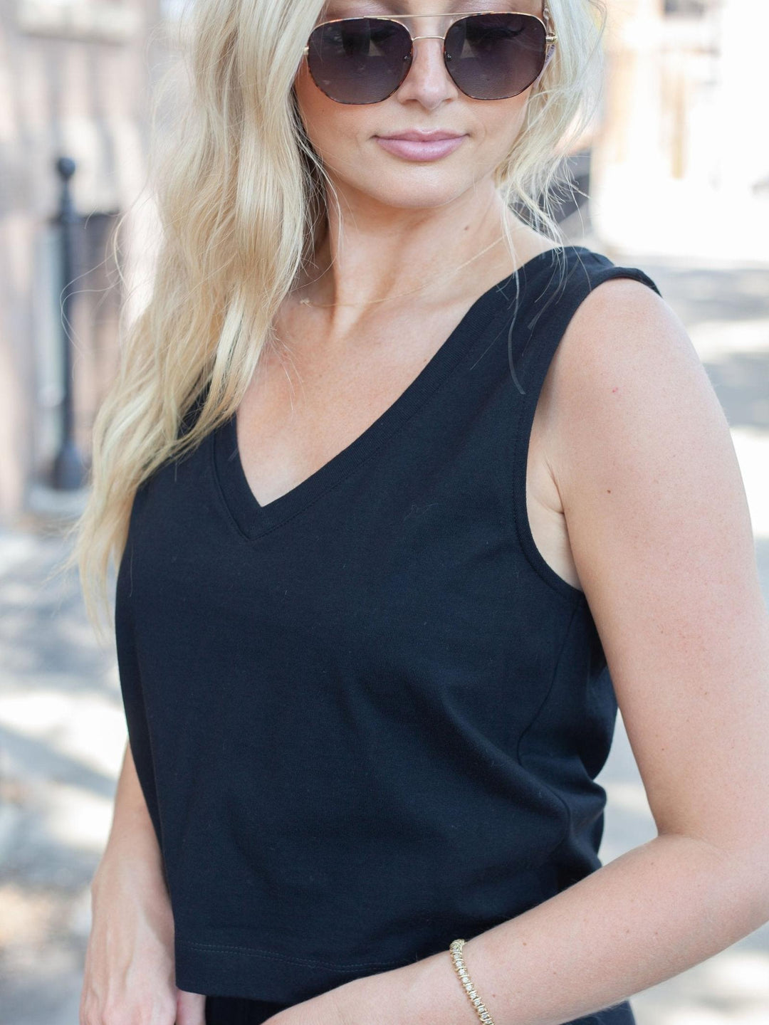 A woman wearing a black sleeveless V-neck top and dark sunglasses, photographed outdoors with a softly blurred street background.