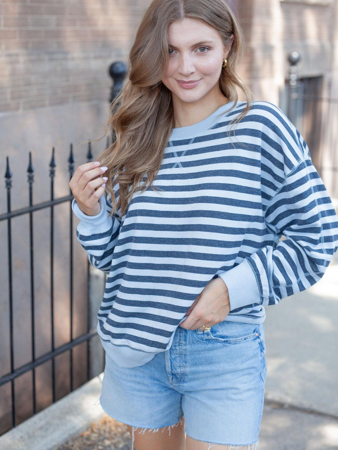A woman wearing a navy blue and white horizontally striped long-sleeve crewneck sweatshirt with light blue denim shorts, standing outdoors in front of a brick wall and black metal fence.