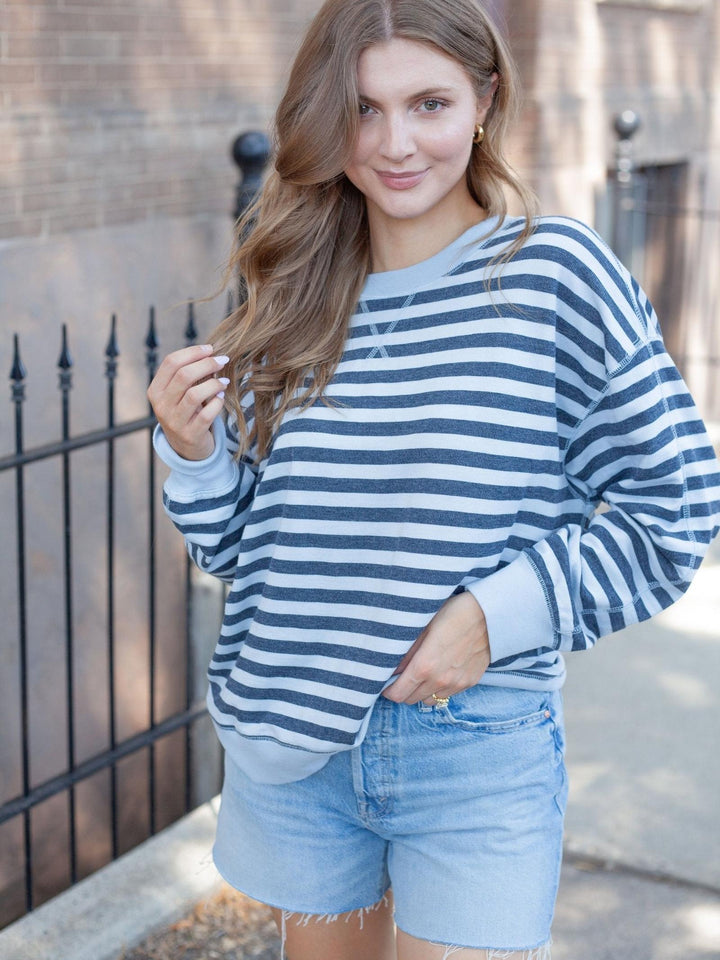 A woman wearing a navy blue and white horizontally striped long-sleeve crewneck sweatshirt with light blue denim shorts, standing outdoors in front of a brick wall and black metal fence.