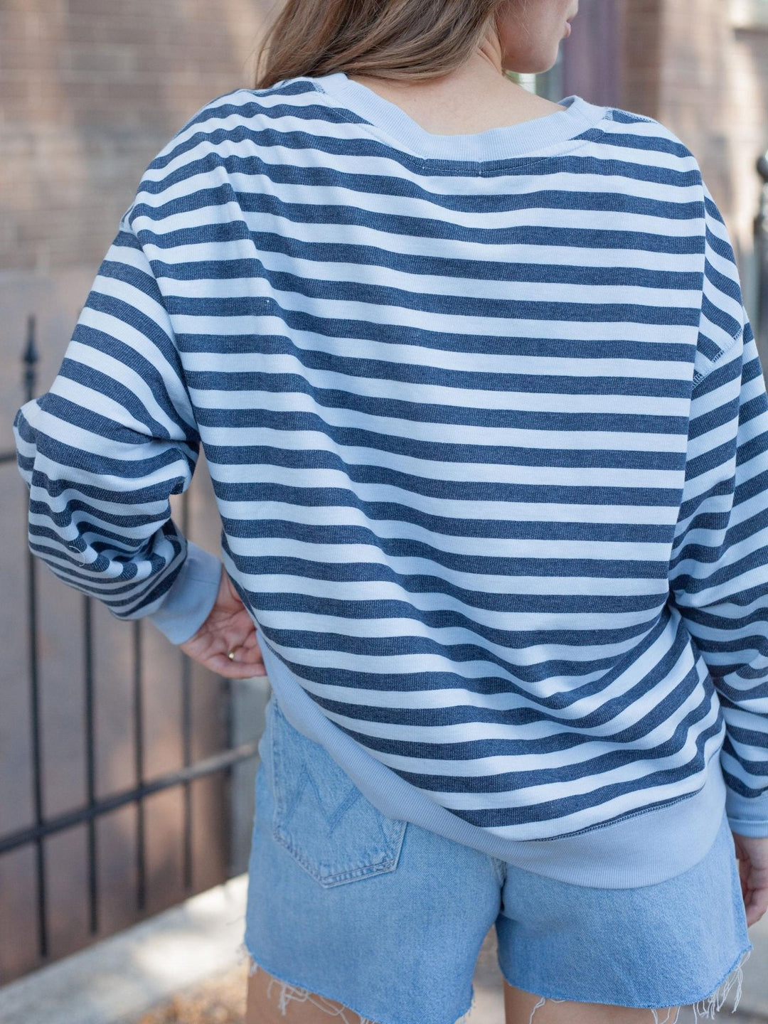 A person shown from the back wearing a blue and white horizontally striped long-sleeve top with light blue denim shorts, standing outdoors against a brick wall and black metal fence.