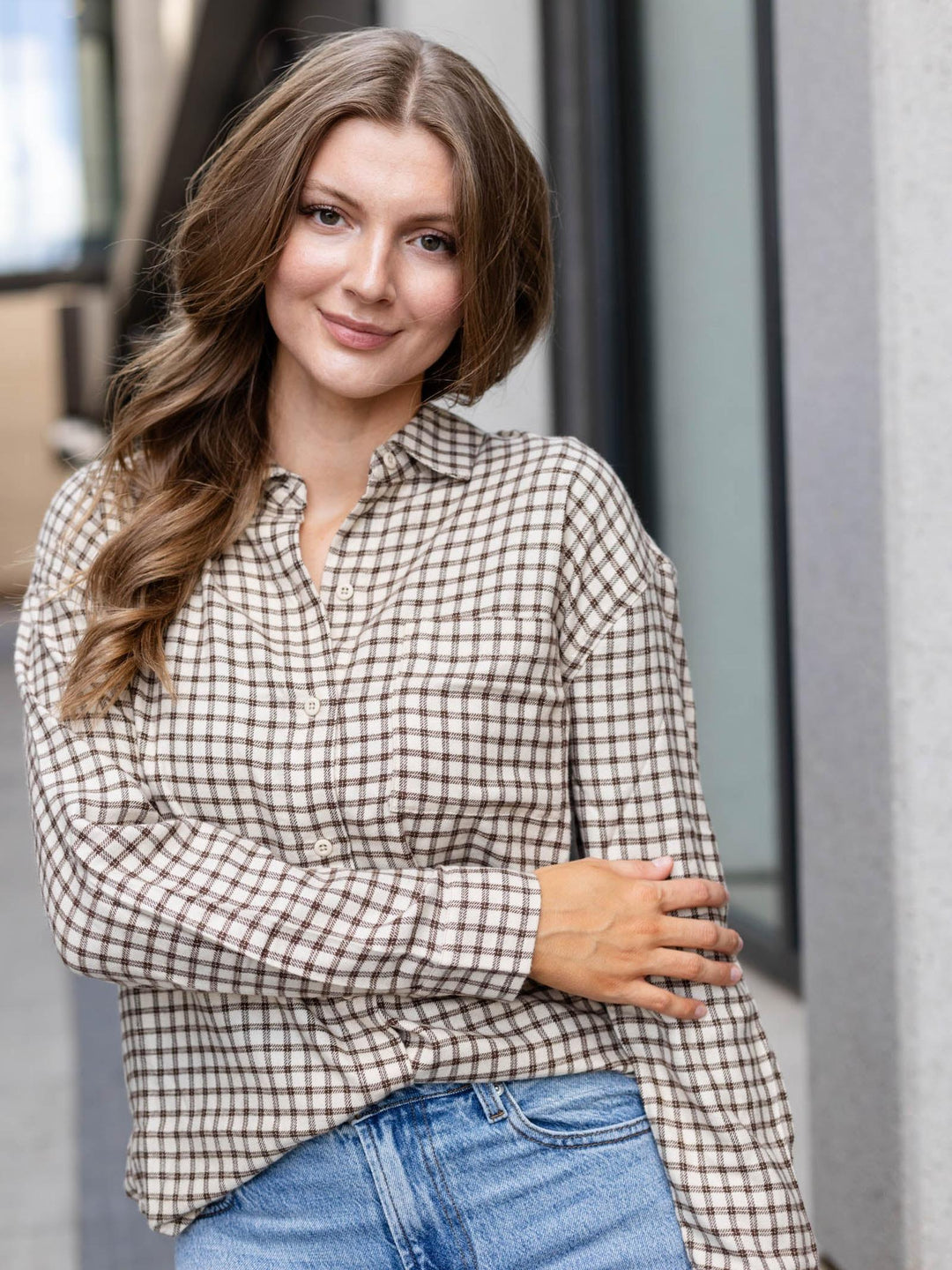 A woman wearing a brown and beige gingham plaid button-down shirt with blue jeans, standing in front of a modern building with gray columns.