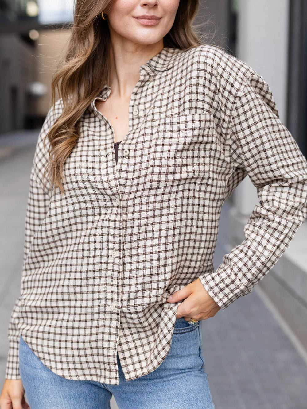 A woman wearing a brown and beige gingham plaid button-down shirt with blue jeans, standing outdoors.