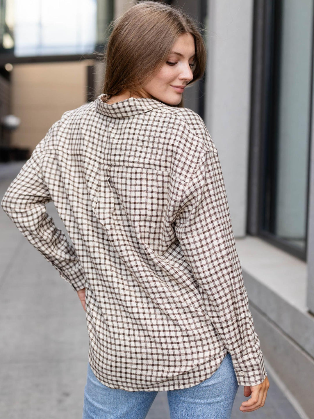 A woman wearing a brown and beige checkered long-sleeve button-up shirt, shown from a rear three-quarter view in an outdoor urban setting.