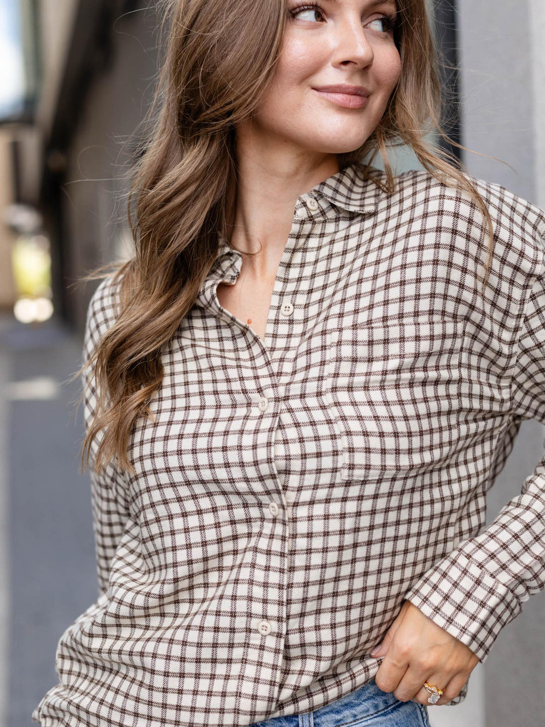 A woman wearing a brown and beige gingham plaid button-down shirt, paired with blue jeans, in an outdoor setting.