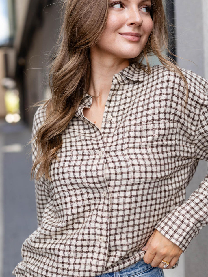 A woman wearing a brown and beige gingham plaid button-down shirt, paired with blue jeans, in an outdoor setting.